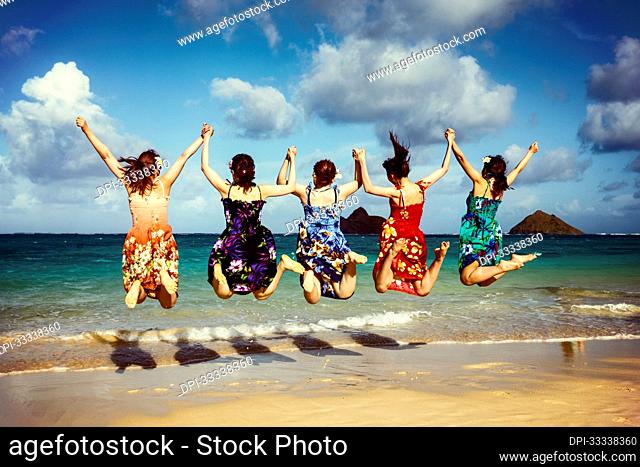 View From Behind Of Japanese Students Jumping In The Air And Holding Hands While On Vacation At Lanikai Beach: Oahu, Hawaii, United States of America