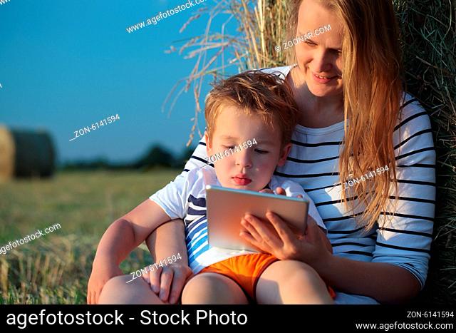 Happy mother with little son on the lap holding a pad and boy playing on it. They sitting by the hay roll, blurry field and sky in background