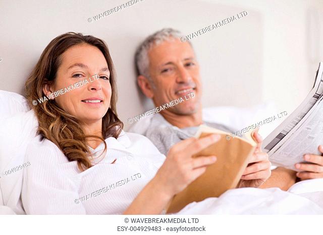 Smiling woman reading a book while her husband is reading the news