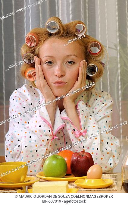 Blonde woman wearing pajamas with curlers in her hair sitting at breakfast table