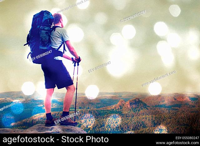 Film grain effect. Tall tourist with poles in hand. Sunny evening in rocky mountains. Hiker with big backpack stand on rocky view point above misty valley
