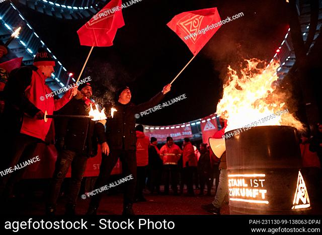 30 November 2023, North Rhine-Westphalia, Duisburg: Participants in an IG Metall action in the course of the current wage dispute wave flags next to a fire...