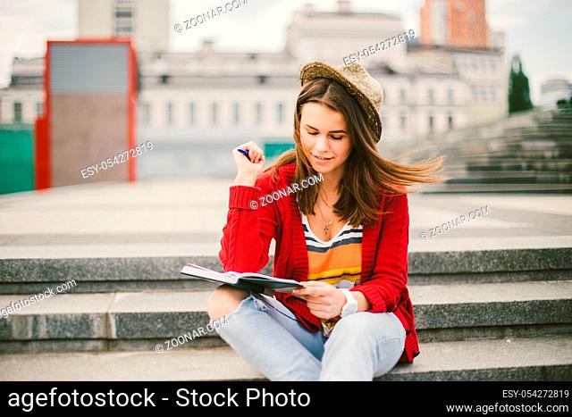 A beautiful, young Caucasian girl sitting on the street smile, the joy, the emotion, sit with notebook and pen in Ruhi. In the red sweater