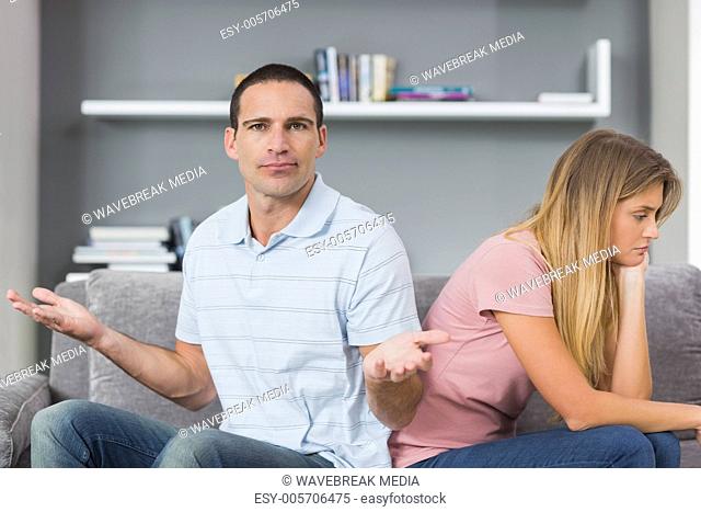 Couple sitting back to back after a fight on the couch with man gesturing at camera