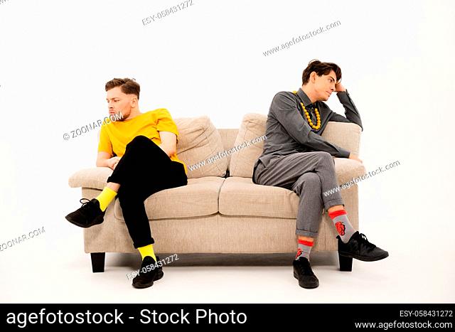 Quarreled guys are sitting on opposite sides of the sofa. Two trendy young mens are sitting on a soft couch and not communicate isolated on white background