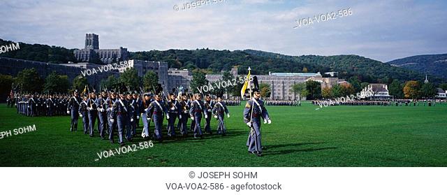 This is the exterior of the West Point Military Academy. Marching are the Homecoming Parade of Cadets in grey uniforms and tall hats, holding long guns