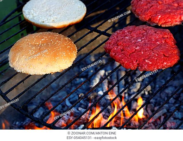 Close up raw beef meat burgers and sesame buns for hamburger cooked on fire barbecue grill with smoke and flame, high angle view