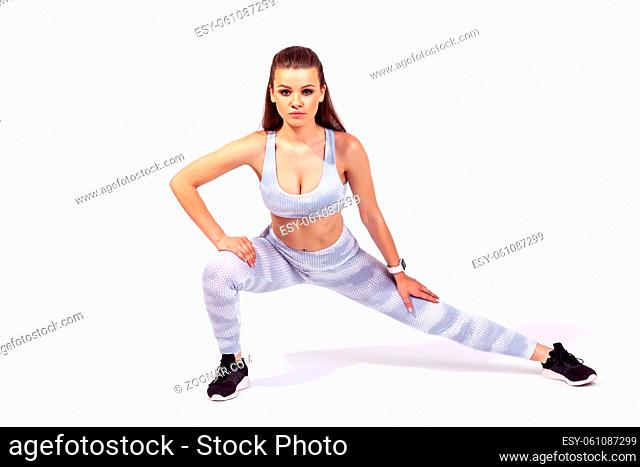 Full length concentrated athletic woman in white sportswear doing lower body sport exercise, stretching legs, warming up muscles for flexibility