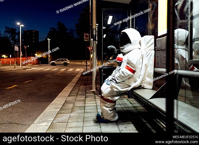 Young female astronaut in space suit waiting at bus stop during night