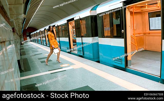 Posing in front of a open doors of an empty subway train, beautiful young girl with long beautiful legs in a yellow spring coat and a white handbag or purse in...