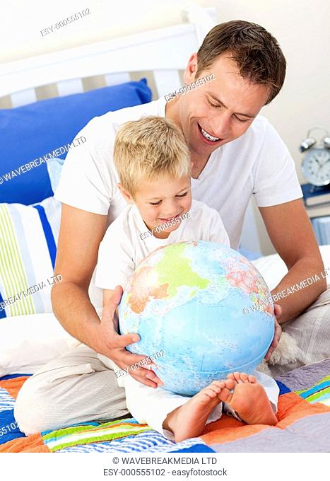 Merry father and his son looking at a terrestrial globe sitting on bed
