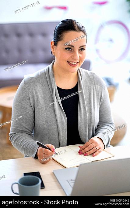 Smiling businesswoman using laptop and writing in diary while working at home