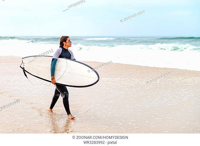 Young female surfer wearing wetsuit, holding surfboard under his arm, walking on beach after morning surfing session