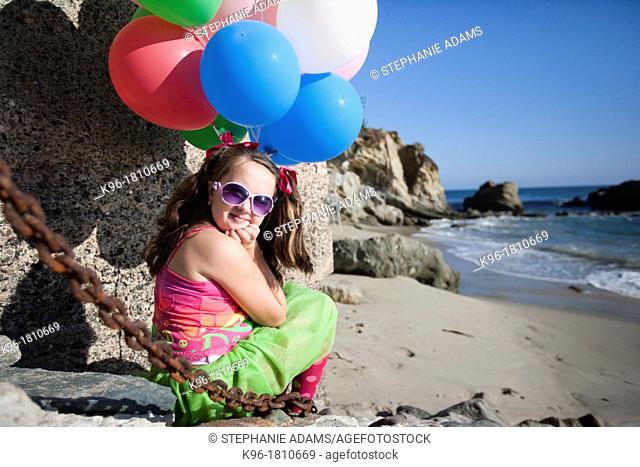 little girl sitting at the ocean with balloons, looking at the camera