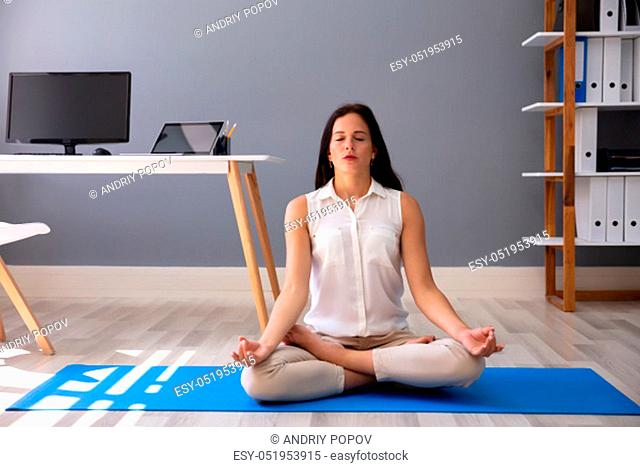 Young Businesswoman Sitting On Blue Exercise Mat Meditating In Office