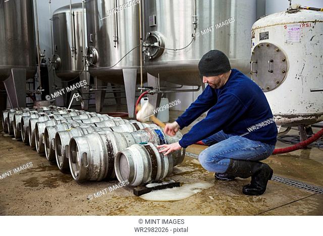 Man kneeling and hammering in a peg into a metal beer keg. Large fermentation tanks