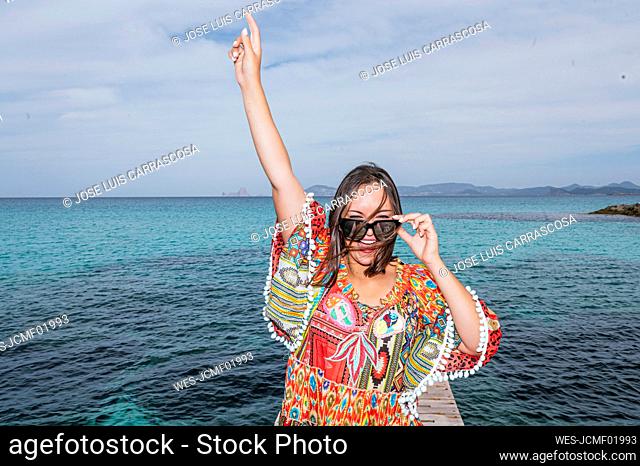 Cheerful woman with hand raised standing in front of water at Formentera island