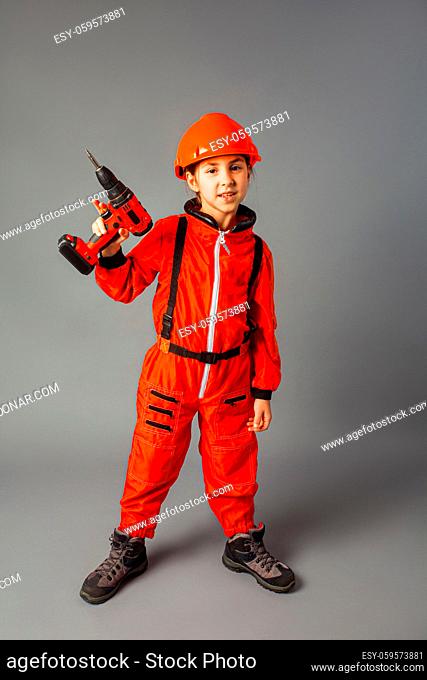 The happy little girl dressed in a red engineering uniform stands on a gray background. She is wearing a protective helmet and holding a screwdriver