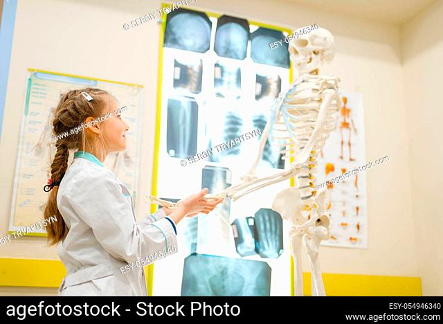 Little girl in uniform playing doctor with human skeleton, playroom. Kid plays medicine worker in imaginary hospital, profession learning, childish dream