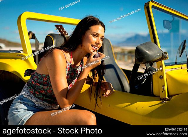 Happy caucasian woman leaning against beach buggy by the sea putting on lipstick