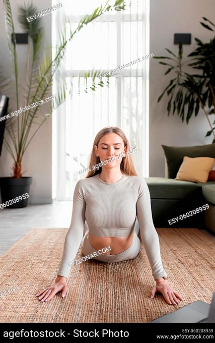 Healthy sport lifestyle concept, blogger girl with closed eyes doing yoga exercises at home in green loft interior watching and repeating trainer exercises and...