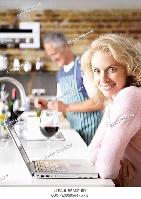 Woman on laptop with wine and man in background preparing salad