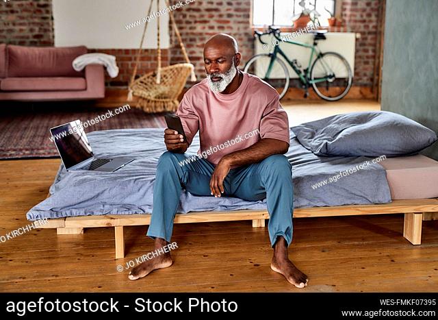 Bald man using smart phone sitting by laptop on bed at home