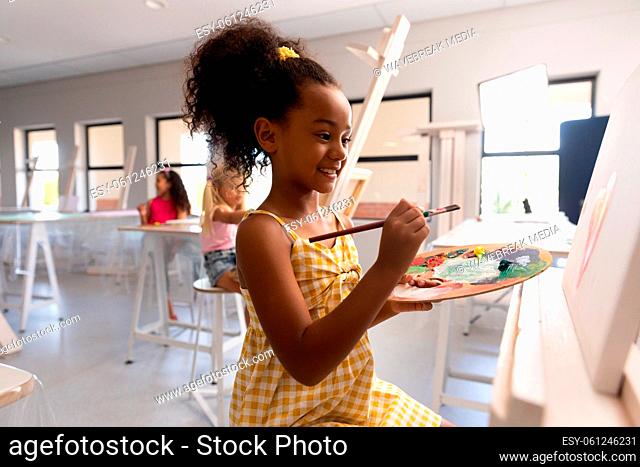 Smiling biracial elementary schoolgirl with palette and paintbrush painting on easel in school