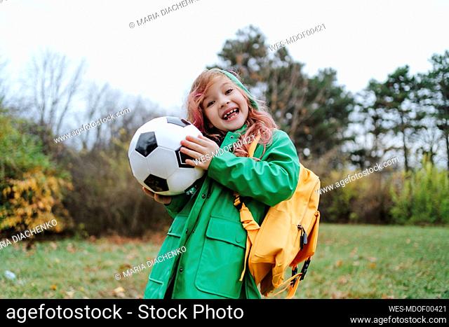 Cheerful girl standing with backpack and soccer ball at park