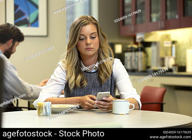 Young Caucasian woman looking at cell phone, while sitting in break area of office with lunch of soup and Hot Tea