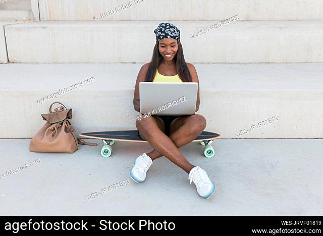 Smiling woman using laptop while sitting cross-legged on skateboard