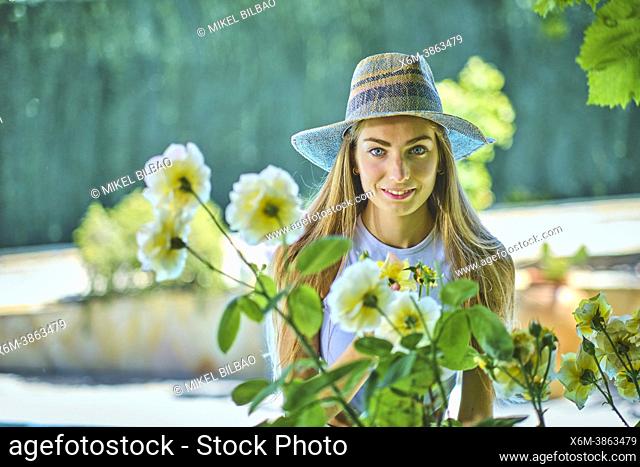 Portrait of a young beautiful caucasian woman in her 20's with long hair and blue eyes wearing a hat and enjoying the rose flowers of a bush outdoor in a garden