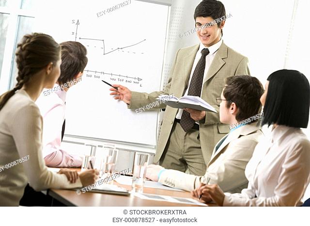 Portrait of handsome businessman pointing at graph on board and looking at his co-workers with smile