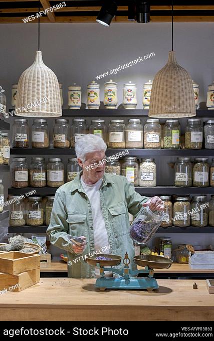 Senior man behind the counter in his shop holding jar of dried lavender blossoms