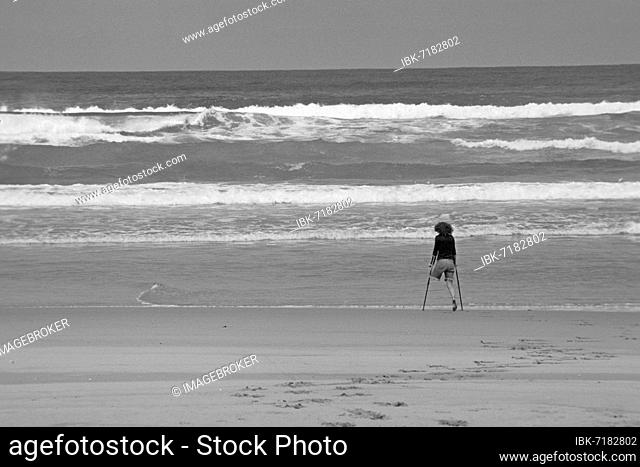 One-legged young woman on crutches by the sea, one-legged, disabled, black and white, black and white shot, disabled on the beach, crutches, walker, walking aid