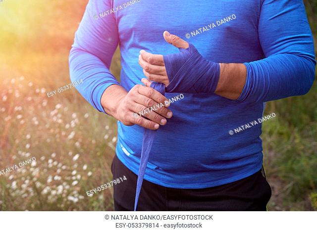 athlete stand and wrap his hands in blue textile elastic bandage before training, sunny summer day, outdoor
