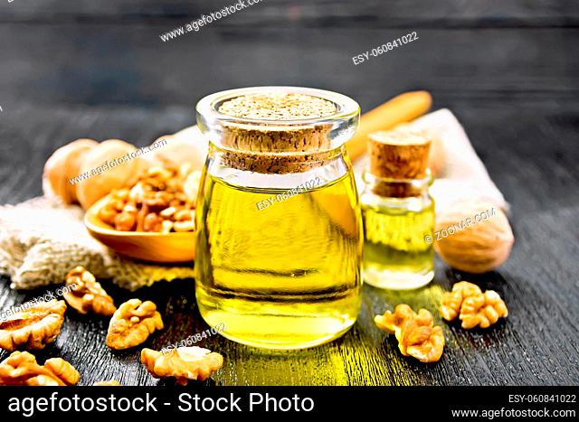 Walnut oil in a jar and a bottle, nuts in spoon and on table, burlap napkin on dark wooden board