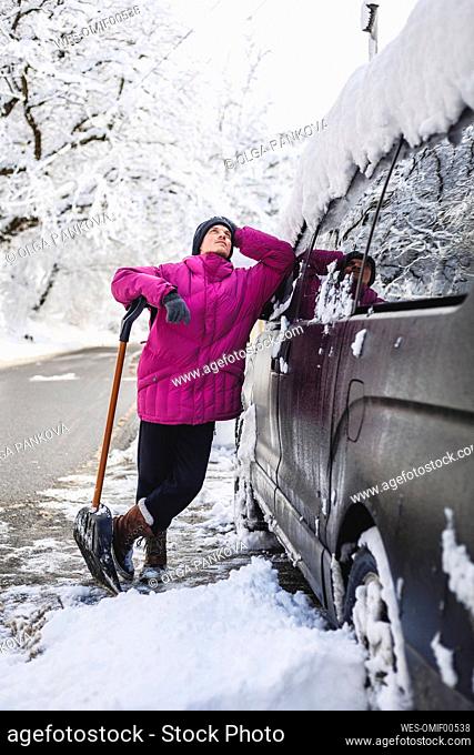 Contemplative man with snow shovel leaning on car