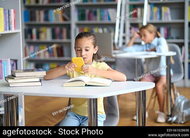 Focused dark-haired teenage girl sitting at the library table and looking at the smartphone screen