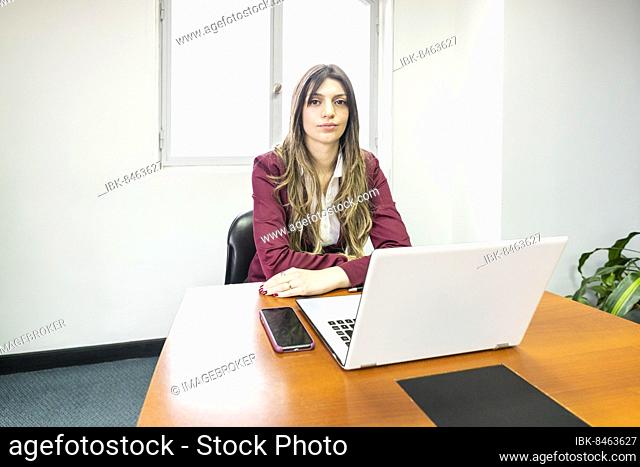 Young blonde business woman sitting at the head of the table in the office meeting room, with her computer and cell phone, looking at the camera