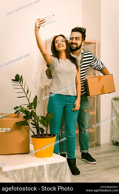 Man and woman in casual clothes standing in new home after relocation, forwarding to new life. Couple making selfie shot in living room witn new furniture
