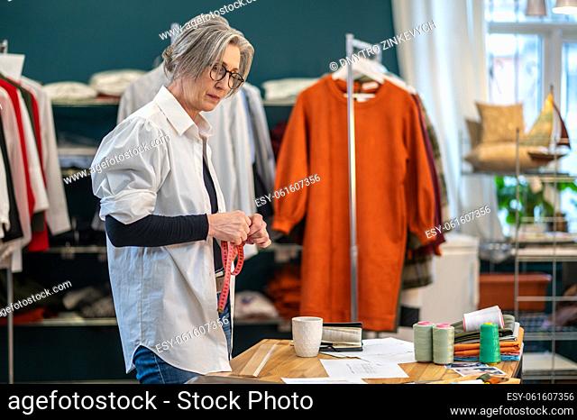 Fashion designer. Gray-haired woman in glasses with measure in hands standing looking at sketch lying on table in sewing workshop