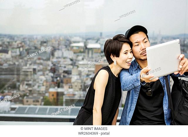 Two people, man and woman taking a selfie with a digital tablet, in front of a view over a large city