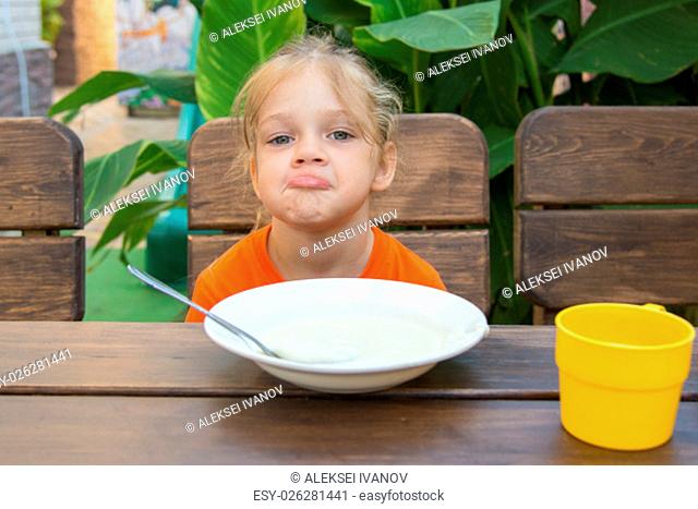 Five-year girl sits at a table with a sour face and did not want to eat porridge for breakfast