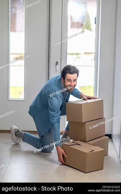 Lucky day. Smiling bearded man looking at camera crouching near boxes gathering them together in hallway of new apartment