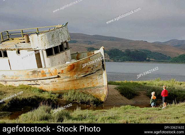Two boys check out an old beached boat along the shore.; Point Reyes, California