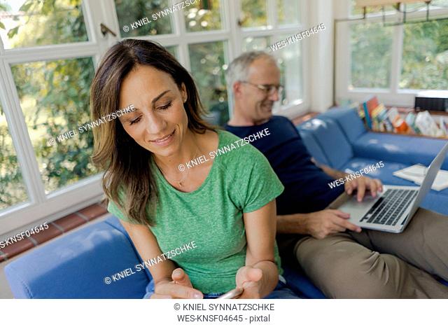 Mature couple sitting on couch at home with woman using cell phone and man using laptop