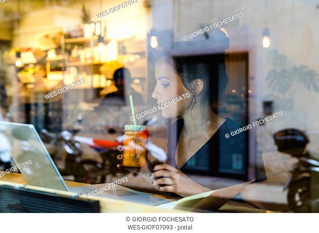 Young woman with smoothie and laptop behind windowpane in a cafe
