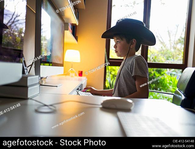 Cute boy in cowboy hat playing video game at computer in home office