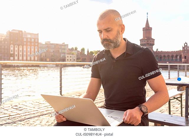 Man using laptop on bridge, river, Oberbaum bridge and buildings in background, Berlin, Germany